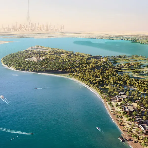 Aerial view of a lush green island with a sandy beach and turquoise water, city skyline in distance.